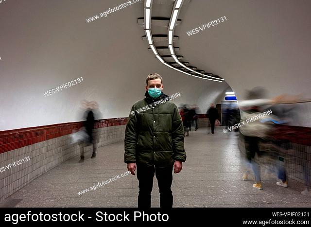 Young man with face mask standing isolated in subway underpass, with people moving around him