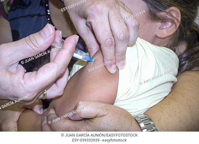 Mature nurse vaccinating a little girl while the mother is hugging her. She is stabbing the needle