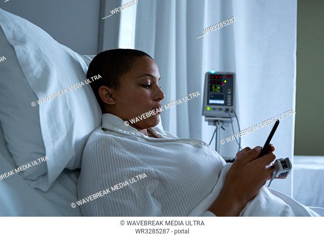 Side view of beautiful mixed race female patient using mobile phone while lying on bed in the ward in hospital