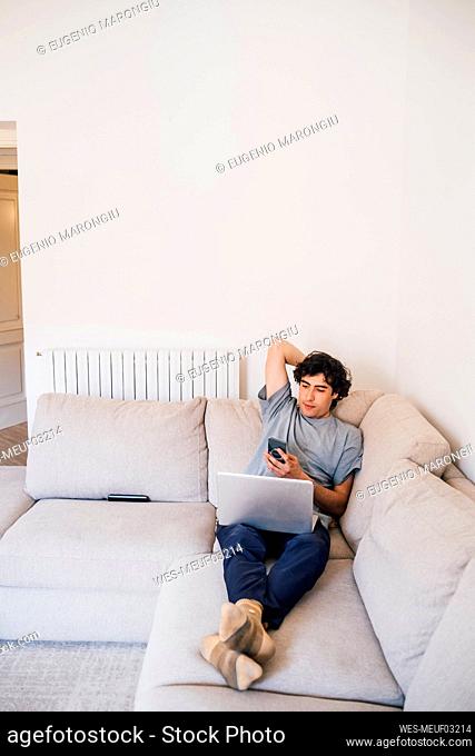Man using mobile phone while sitting with laptop on sofa in living room