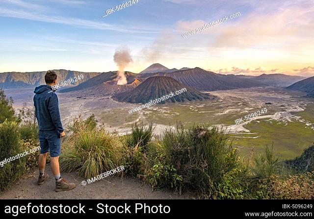 Young man in front of volcanic landscape at sunset, view in Tengger Caldera, smoking volcano Gunung Bromo, in front Mt. Batok, in the back Mt