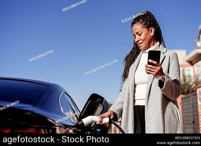 Smiling woman standing with smart phone and charging car on sunny day