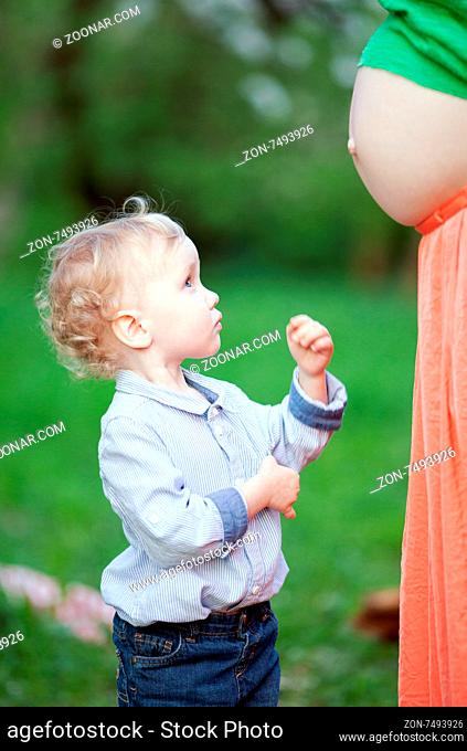 Lovely little boy looking at pregnant mothers belly outdoor. He is expecting to have brother or sister