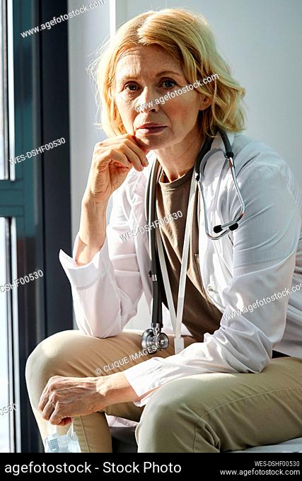 Mature doctor with hand on chin sitting in hospital