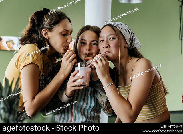 Teenage girls standing in kitchen sharing fresh fruit smoothie with drinking straws