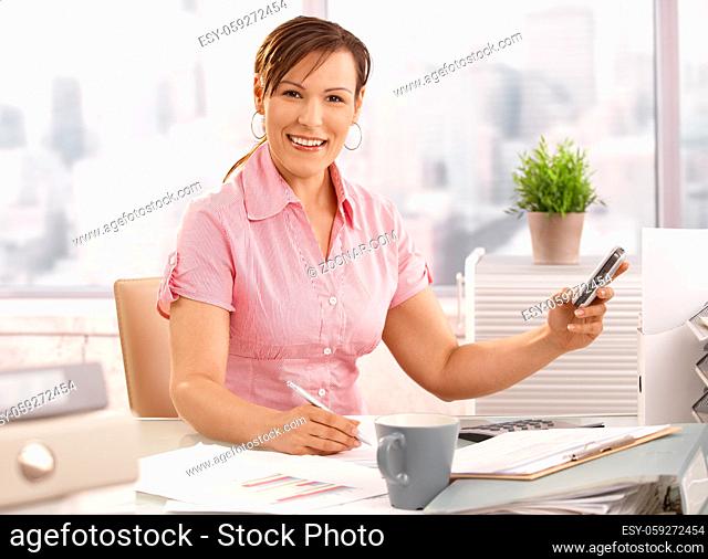 Young businesswoman sitting at office using mobile phone, smiling