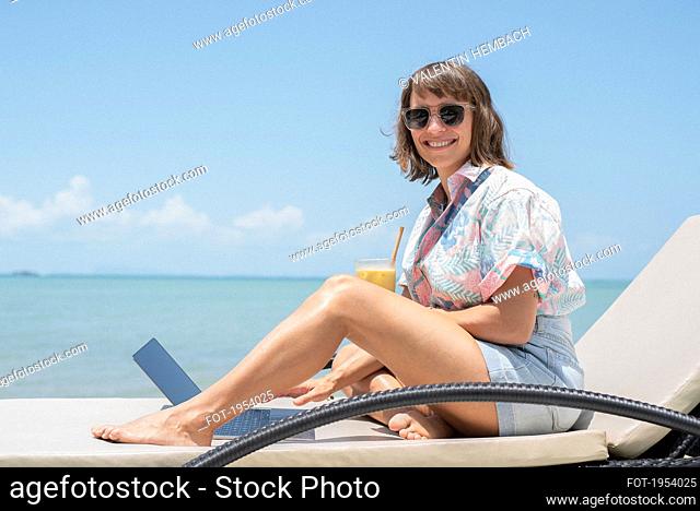 Portrait happy woman with cocktail and laptop on sunny beach lounge chair