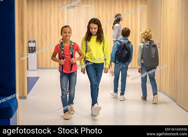 School day. Joyful girls with backpacks walking and boys behind with their backs to camera in illuminated corridor of school