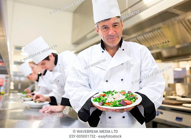 Cheerful chef presenting his salad with others preparing salads in kitchen