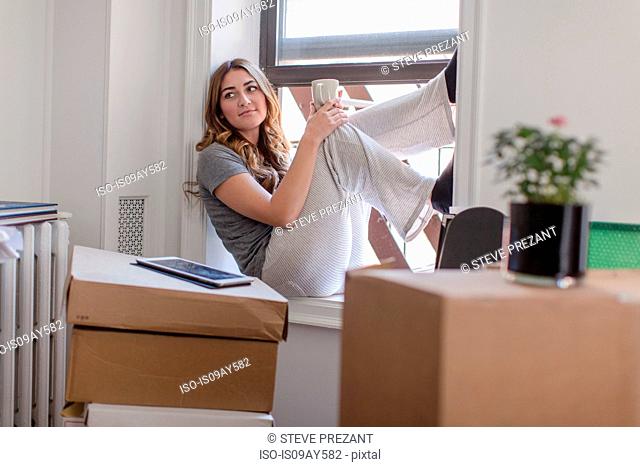 Young woman sitting on windowsill of new home, holding hot drink, cardboard boxes in room
