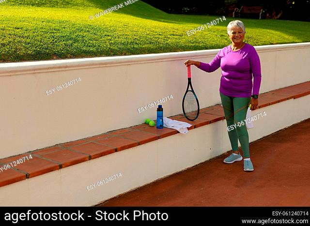 Portrait of happy biracial senior woman with racket, ball, water bottle and towel on bench at court