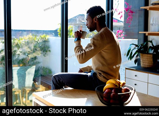 Happy african american man sitting at countertop in kitchen, drinking coffee