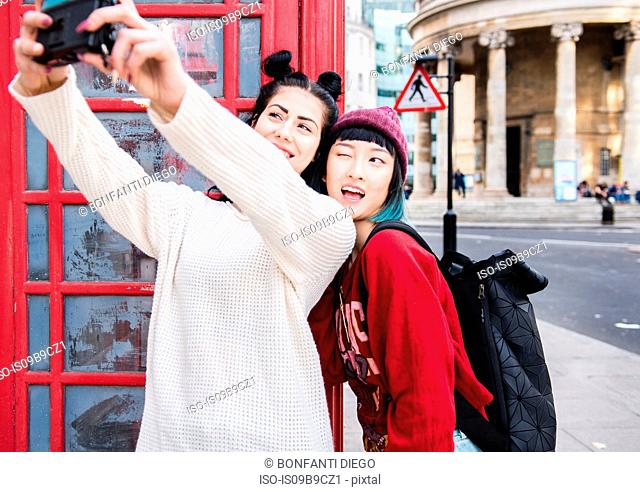 Two young stylish women taking selfie by red phone box, London, UK