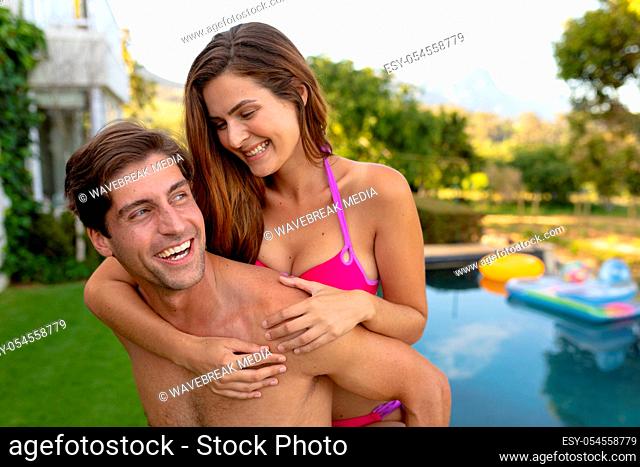 Front view of a Caucasian couple wearing beachwear in a garden, the man carrying the woman piggyback, standing beside a swimming pool