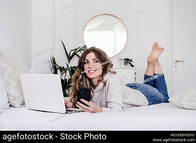 Thoughtful beautiful woman with laptop and smart phone lying on bed
