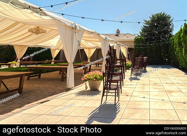 Garden wedding venue with stacks of wooden chairs outside a pavilion with canopy. Chandelier, wooden tables, and string of bulbs can also be seen in this...