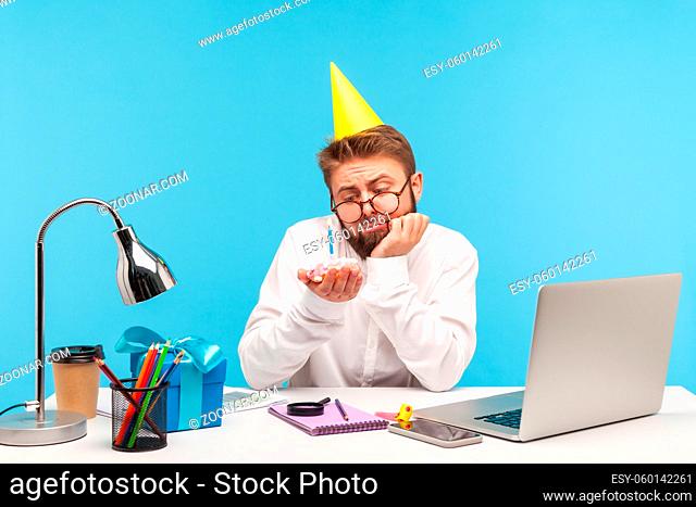 Bored unhappy man office worker in eyeglasses and cone hat looking at cake with candle, lonely celebrating birthday sitting at workplace, remote work