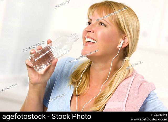 Pretty blonde woman with towel and ear phones drinking from water bottle in her kitchen