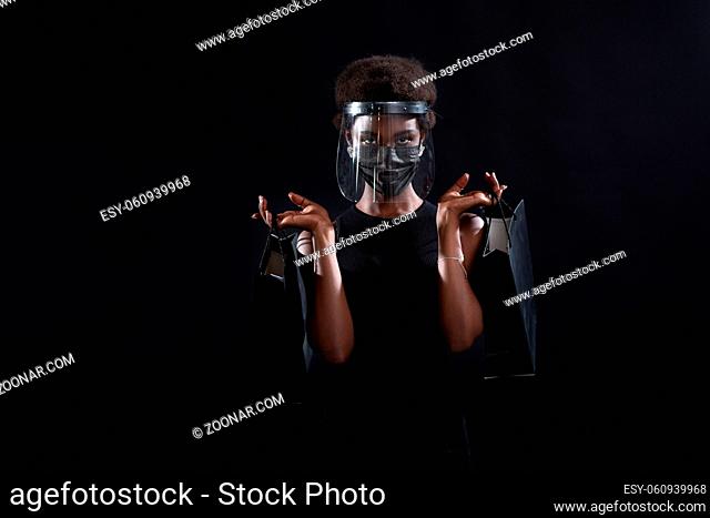 African american woman in black medical face mask and transparent protective face shield holds black shopping bags. Safety on Black Friday concept