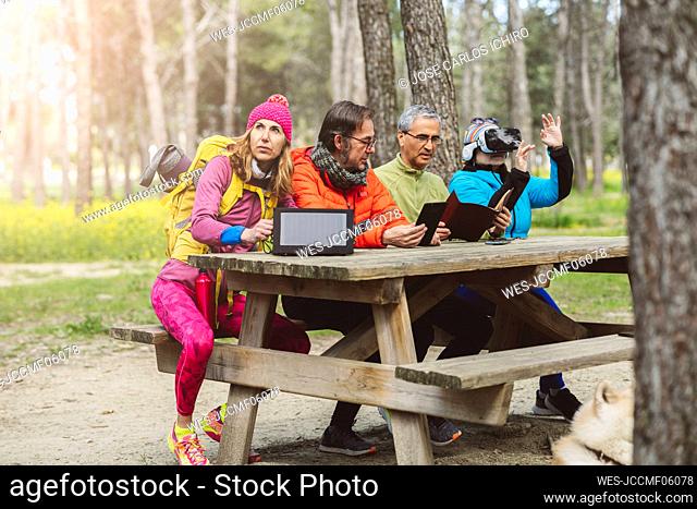 Mature man with solar panel sitting by friends at picnic table in forest