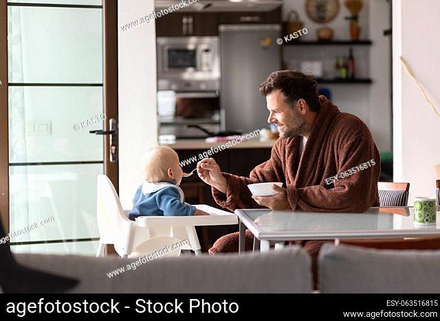 Father wearing bathrope spoon feeding hir infant baby boy child sitting in high chair at the dining table in kitchen at home in the morning