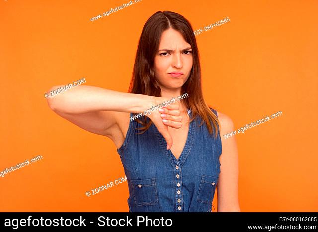 Dislike. Naughty dissatisfied brunette woman in denim dress frowning angrily and showing thumbs down gesture, expressing disapproval, negative sign