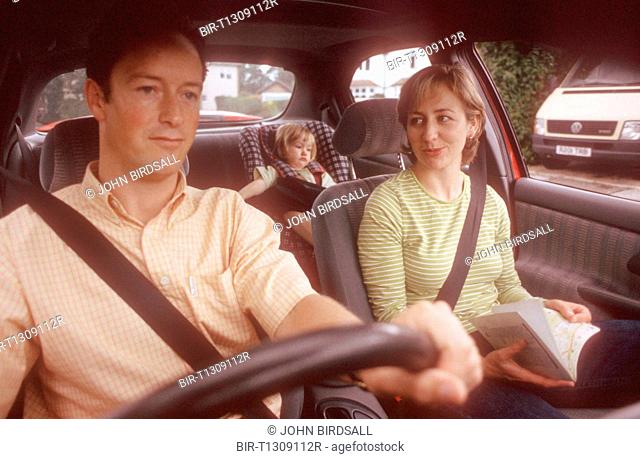 Family sitting in car with father driving, mother map reading and young child strapped into car seat