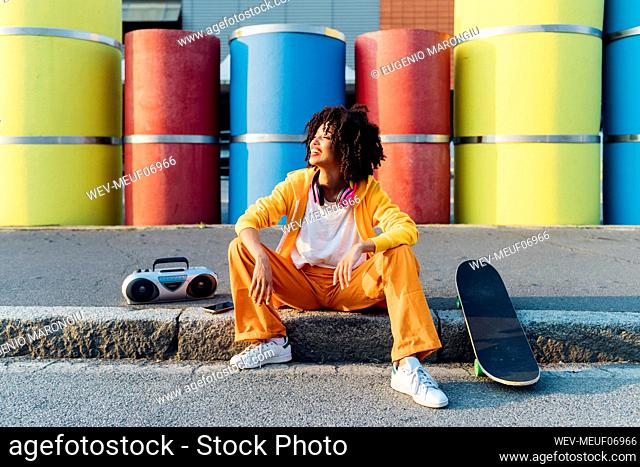Happy young woman with skateboard and boom box sitting in front of multi colored pipes