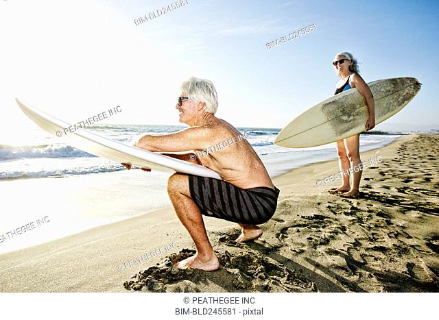 Older Caucasian couple standing on beach holding surfboards