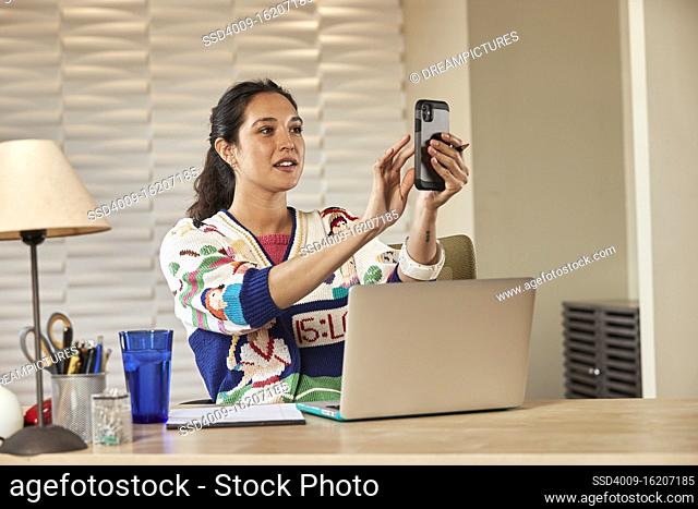 Young ethnic woman sitting at desk with laptop computer taking notes on notepad and using smartphone