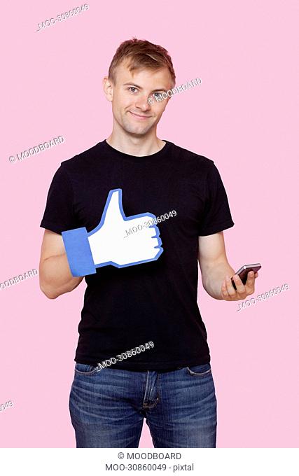 Portrait of a happy young man with cell phone holding fake like button over pink background