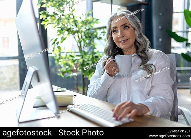 Working place. Good-looking business woman sitting at the comeputer and having tea