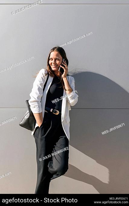 Smiling businesswoman with hand in pocket talking on mobile phone in front of wall