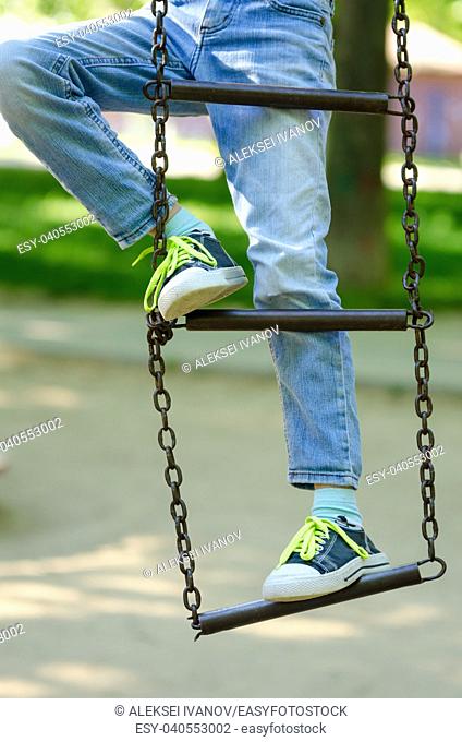 Legs of a child in sneakers with light green laces on a metal ladder on chains