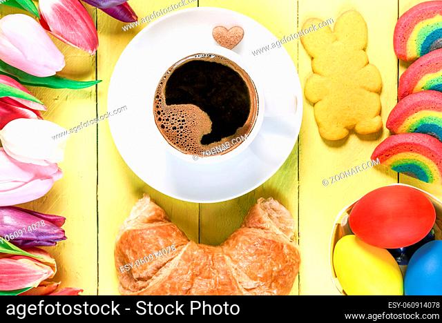 Easter morning with a cup of coffee, rainbow cookies, painted eggs, bunny shaped cookie, croissant and multicolored tulips, on a yellow wooden table