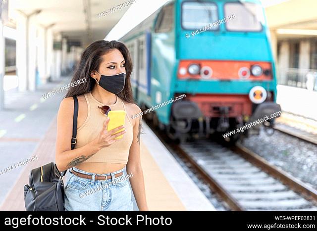 Young woman wearing face mask looking away while using mobile phone standing at station