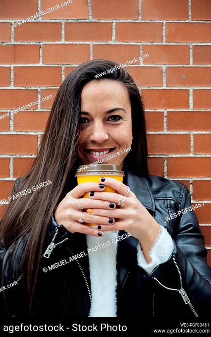 Smiling woman in leather jacket holding reusable cup against brick wall