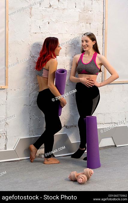 Beautiful young women in sports out fits talking standing next to the wall holding a yoga mat, smiling to each other, standing on a white brick wall background