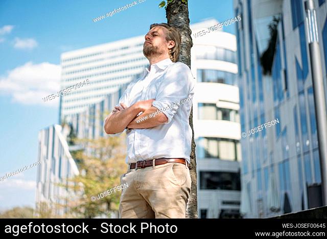 Businessman having a break in the city leaning against a tree trunk