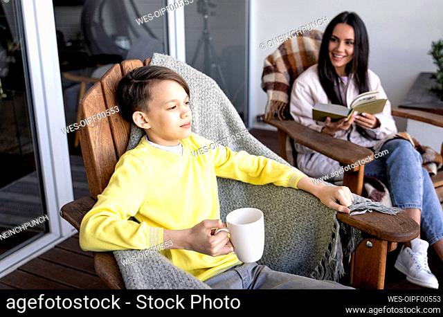 Boy with mug sitting with mother on chair on balcony
