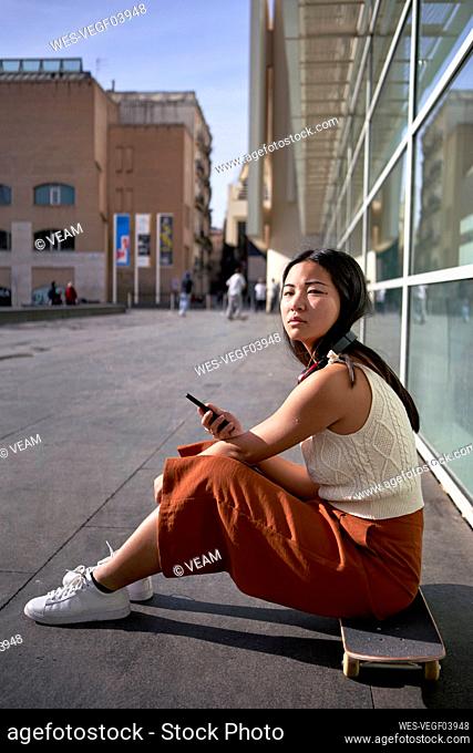 Young woman with smart phone sitting on skateboard during sunny day