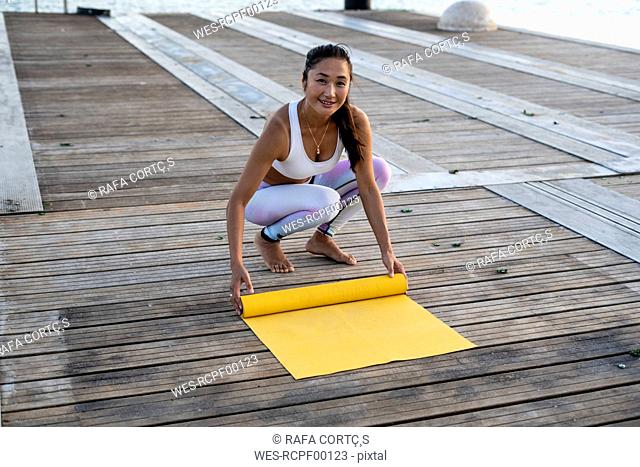 Asian woman practicing yoga on a pier at harbour, rolling out her yellow yoga mat