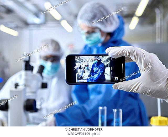 Hand holding smartphone, photographing chemists, working in industrial laboratory