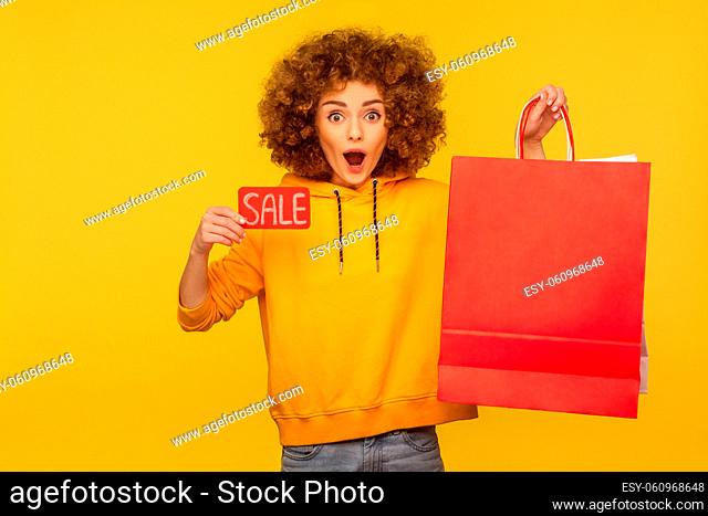 Portrait of shocked surprised curly-haired hipster girl in hoodie holding shopping bags with blank area for advertising text and showing Sale inscription