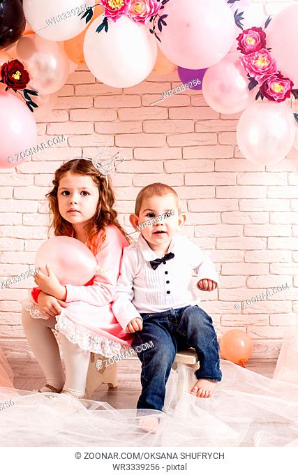 Boy and girl with crowns under birthday balloon and paper flower arch decorations. Childish photozone for celebration