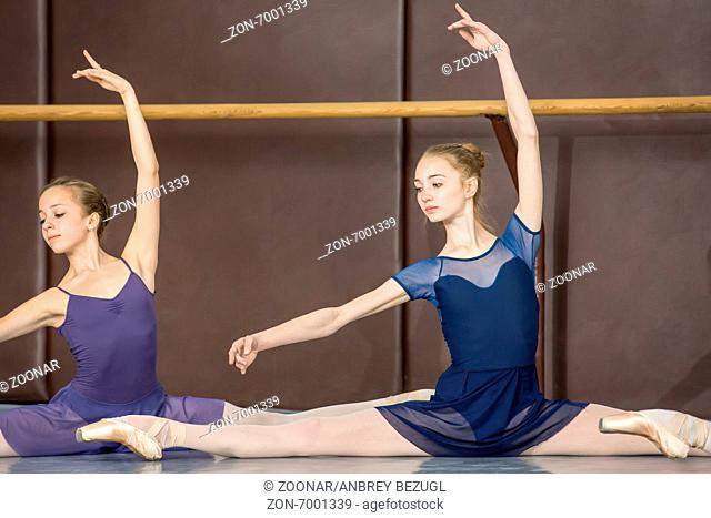 Young ballerinas on the lesson of classical choreography sitting in the splits gracefully raising his hands up near barre