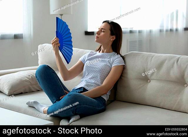 Woman wear casual clothes sitting resting on couch in living room suffers from unbearable hot summer day weather waving hand blue fan reduces heat cooling...