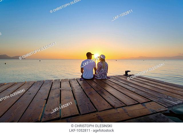Young couple sitting at the end of a jetty in the morning atmosphere and enjoying the view of the sunrise. Playa de Muro beach, Alcudia, Mallorca