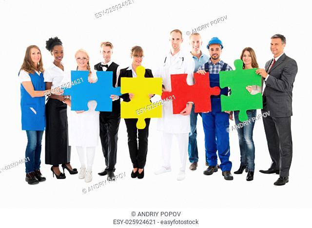 Portrait of confident people with various occupations holding jigsaw pieces while standing against white background