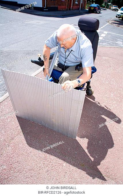 Male wheelchair user setting up a portable folding travel ramp to use at a roadside kerb